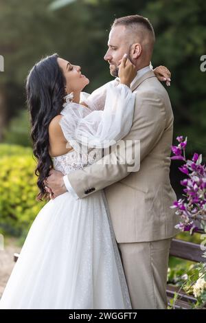 Hochzeitsporträt lächelnder Jungvermählter. Ein stilvoller Bräutigam in einem beigefarbenen Anzug und eine süße Brünette in einem weißen Kleid sind zärtlich im Park. Stockfoto