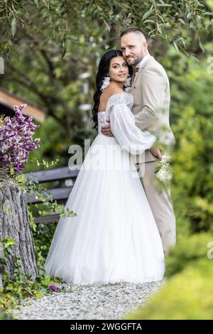 Hochzeitsporträt lächelnder Jungvermählter. Ein stilvoller Bräutigam in einem beigefarbenen Anzug und eine süße Brünette in einem weißen Kleid sind zärtlich im Park. Stockfoto