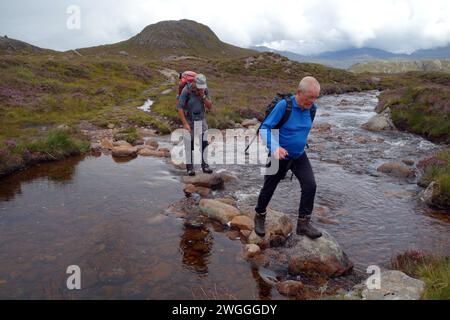 Two Men (Wanderer) auf Stepping Stones from the Outflow of 'Loch na h-Oidhche' im Flowerdale Forest, Wester Ross, North West Highlands, Schottland, Großbritannien Stockfoto