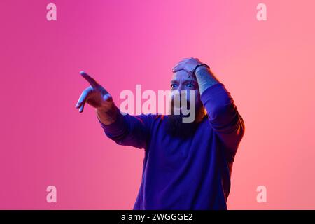 Wow. Bärtiger Glatzkopf in seinen 30ern, stehend mit weit geöffneten Augen, Mund, schockiertem Gesicht und mit dem Finger auf rosa Hintergrund in Neon Stockfoto