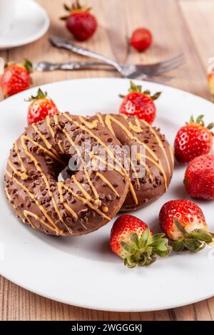 Das Bild zeigt zwei Schokoladen-Donuts auf einem weißen Teller, der auf einem Holzsockel steht. Die Donuts werden mit frischen Erdbeeren und Kuchengabeln nex garniert Stockfoto