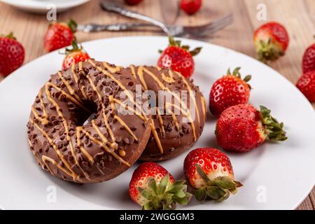 Das Bild zeigt zwei Schokoladen-Donuts auf einem weißen Teller, der auf einem Holzsockel steht. Die Donuts werden mit frischen Erdbeeren garniert und mit ca. Stockfoto