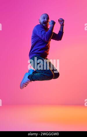 Das Bild eines bärtigen Glatzen-Mannes in lässiger Kleidung, der emotional im Neonlicht vor rosa Hintergrund springt Stockfoto