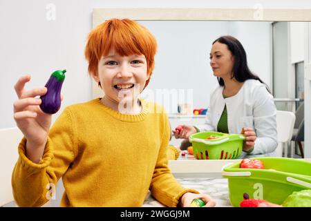 Rothaariger Junge, der SpielzeugAuberginen beim Sprachtraining mit seinem Logopäden hält. Sprach- und Sprachtherapie der Kinder Stockfoto