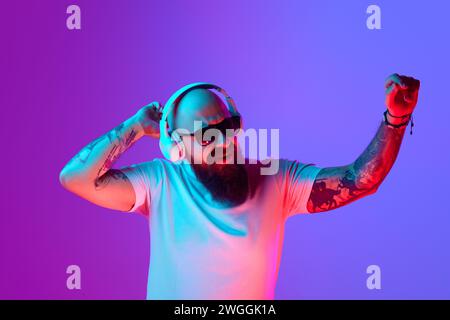DJ. Bärtiger, künstlerischer Glatze Mann mit Tätowierungen, in weißem T-Shirt und Sonnenbrille, die vor verlaufendem lila Hintergrund im Neonlicht steht Stockfoto