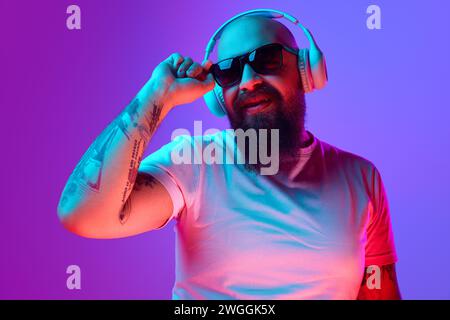 DJ. Bärtiger, künstlerischer Glatze Mann mit Tätowierungen, in weißem T-Shirt und Sonnenbrille, die vor verlaufendem lila Hintergrund im Neonlicht steht Stockfoto