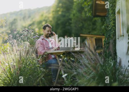 Mann, der im Garten am Laptop arbeitet, telefoniert. Geschäftsmann, der aus der Ferne vom Heimbüro im Freien arbeitet und über neue Geschäfte oder kreative Ideen nachdenkt Stockfoto