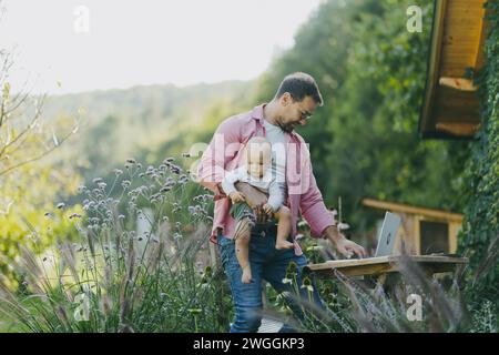 Vater hält Baby in der Hand, während er im Garten am Laptop arbeitet. Geschäftsmann, der aus der Ferne vom Home Office im Freien arbeitet und sich um den kleinen Sohn kümmert. Lebensarbeit Stockfoto