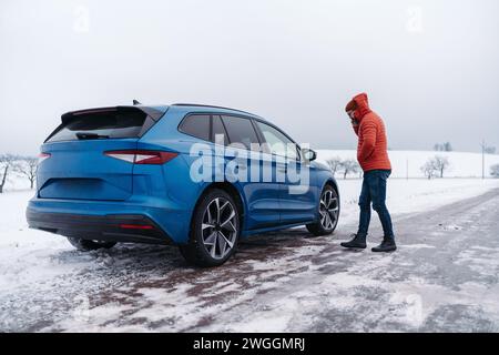 Wütender Mann, der am Elektroauto steht, die Batterie hat keine Energie mehr, bevor er das Ziel erreicht. Ein Mann ruft Hilfe an und wartet auf einen Pannendienst Stockfoto