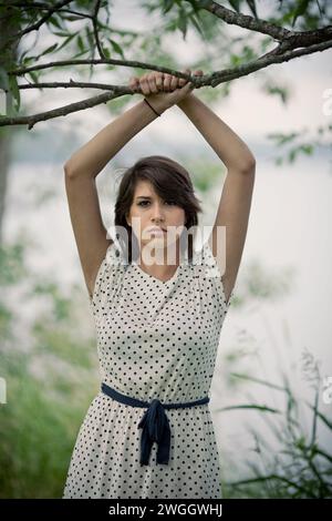 Eine junge Frau mit weißem Kleid und einem intensiven Blick posiert für ein Porträt neben einem Baum. Stockfoto