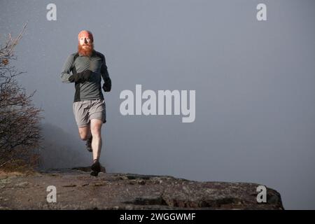 Ein Trailläufer mit langem Bart läuft am Rand einer Klippe im Schnee und Nebel entlang. Stockfoto