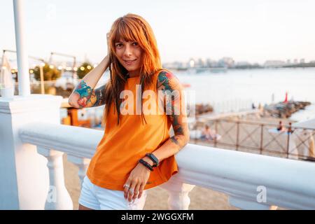 Porträt einer jungen, lächelnden, schönen kaukasischen Frau mit Tätowierungen, die sich auf die Balustrade stützen und in die Kamera schauen. Freiheitsbegriff und Psychologie. Stockfoto