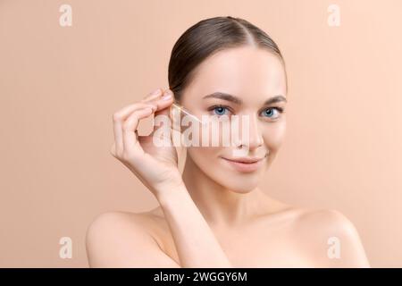 Schöne junge Frau, die im Studio Serum auf beigefarbenem Hintergrund auf ihr Gesicht aufträgt Stockfoto