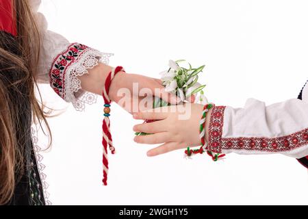 Bulgarische Kinder Jungen und Mädchen in traditionellen Folklore-Kostümen mit Frühlingsblumen, Schneetropfen und handgefertigten Wollarmband Martenitsa Symbol von Baba Marta Stockfoto