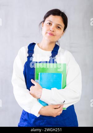 Junge Mechanikerin in blauen Overalls, die Arbeitsbücher hält Stockfoto