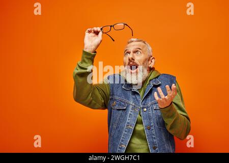 Gut aussehender, überraschter reifer Mann in lebendiger Kleidung, der eine Brille hält und vor orangefarbenem Hintergrund posiert Stockfoto