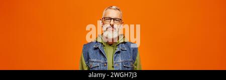 Funky reifer Mann in lebendigem Hoodie und stylischer Weste mit Blick auf die Kamera vor orangefarbenem Hintergrund, Banner Stockfoto