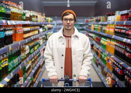 Männlicher Käufer mit Gläsern und Beanie, der einen Wagen in einem Getränkehaus hält. Porträt eines Mannes mit einem Einkaufswagen in einem Supermarkt, der die Kamera vor dem Hintergrund von Regalen mit kohlensäurehaltigen Getränken und Säften betrachtet Stockfoto