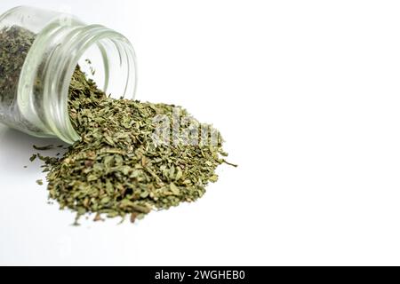 Grünes getrocknetes Kraut, das aus einer Glasflasche oder einem Glas verschüttet wird, weicher Fokus in Nahaufnahme, isoliert auf weiß Stockfoto