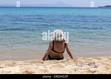 Ältere Frau in Badekleidung, die auf Sand am Meer sitzt Stockfoto