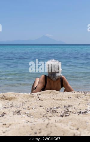 Ältere Dame in Badekleidung, die auf Sand am Meer sitzt Stockfoto