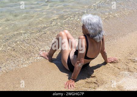 Alte Frau in Badekleidung, die auf Sand am Meer sitzt Stockfoto