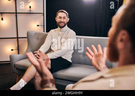 Fröhliche bärtige Männer in lässigem Outfit lächeln und seinen Interviewer anschauen, während sie im Studio sitzen Stockfoto