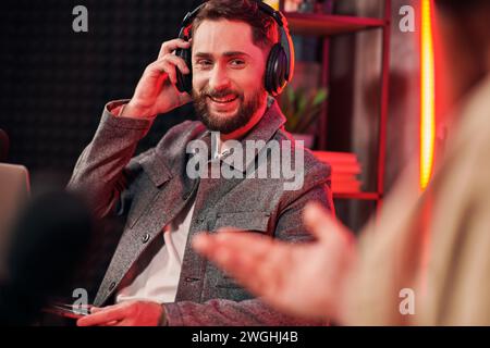 Gutaussehender, fröhlicher Mann in lässiger, gemütlicher Kleidung, der mit Laptop im Studio neben seinem Podcaster sitzt Stockfoto