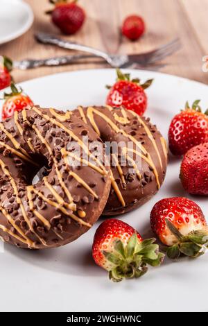 Das Bild zeigt zwei Schokoladenkrapfen auf Einer weißen Platte, die auf Einer Holzfläche thront. Die Donuts Werden Mit Frischen Erdbeeren Und Arrang Garniert Stockfoto