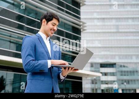 Ein lächelnder hispanischer Geschäftsmann steht mit einem offenen Laptop vor einem modernen Büro und verkörpert das Konzept der zugänglichen Technologie und Geschäftseffizienz in der heutigen schnelllebigen Welt. Stockfoto