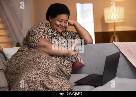 Seitenansicht Porträt einer plumpen schwarzen Seniorin, die zum Videochat winkt, auf der Couch in einem gemütlichen Zuhause sitzt und glücklich lächelnd im Kopierraum sitzt Stockfoto