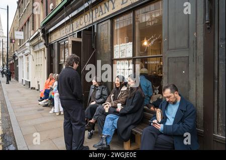 Gruppe von Leuten, einige auf Holzbank, vor Verde & Company Teestube und Coffee Shop in der Brushfield Street London UK Stockfoto