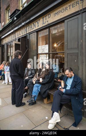 Gruppe von Leuten, einige auf Holzbank, vor Verde & Company Teestube und Coffee Shop in der Brushfield Street London UK Stockfoto