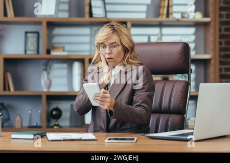 Eine besorgte Geschäftsfrau sitzt am Bürotisch und schaut schockiert auf den Bildschirm des Mobiltelefons, das sie in ihren Händen hält. Hält Hand an Gesicht und liest Nachricht. Stockfoto