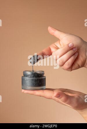 Weibliche Hände halten ein Glas mit schwarzer kosmetischer Gesichtspflege und tragen es auf den Finger auf braunem Hintergrund auf. Kosmetische Industrie. Stockfoto