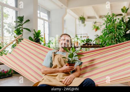 Langhaariger blonder kaukasischer Gärtner, der sich in der Hängematte entspannt und Ficus im Topf im Gartencenter hält. Stockfoto