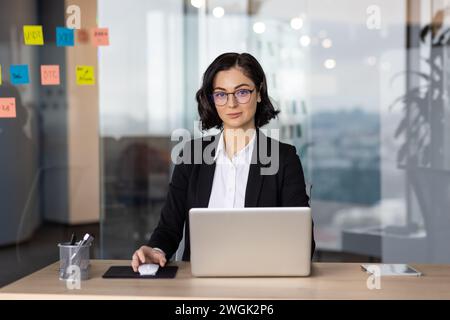 Kaukasische Frau in Brille mit Kurzhaarschnitt am Schreibtisch sitzend und Hand auf Computermaus in Glaswand-Büro. Perspektive für den Mitarbeiter des Unternehmens, der einen neuen Tag im persönlichen Arbeitsbereich beginnt. Stockfoto