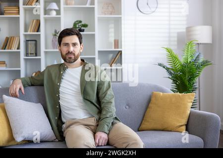 Selbstbewusster Brünette Mann in lässigem Outfit, der sich auf der Rückseite des Sofas mit einem Biegearm im stilvollen Innenraum lehnt. Hübscher Apartmentbesitzer, der sich mit der Investition in neues Wohnzimmerdesign zu Hause zufrieden fühlt. Stockfoto