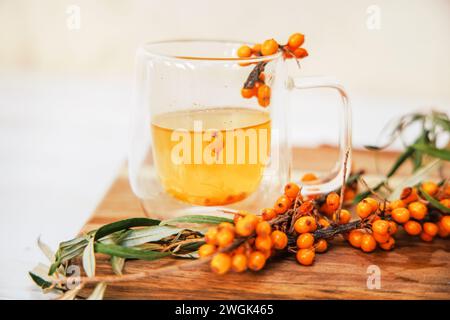 Tasse mit duftendem Vitamintee aus Sanddorn auf einem Holztisch. Frische Sanddornbeeren auf einem Zweig sind bereit für die Vorbereitung einer Heilung Stockfoto