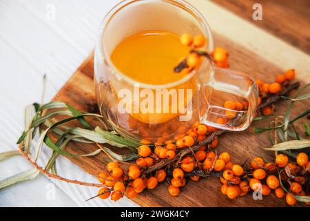 Tasse mit duftendem Vitamintee aus Sanddorn auf einem Holztisch. Frische Sanddornbeeren auf einem Zweig sind bereit für die Vorbereitung einer Heilung Stockfoto