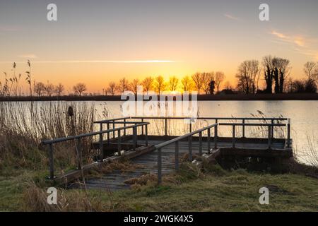 Der Steg scheint verlassen zu sein und wartet vielleicht auf ein paar Fischer. Das Schilf an der Seite und die Bäume im Hintergrund verleihen diesem Foto eine ländliche Atmosphäre Stockfoto