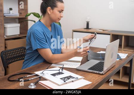 Ärztin in blauen Peelings analysiert die Ergebnisse des Ultraschallpatienten sorgfältig und macht Notizen auf dem Laptop Stockfoto