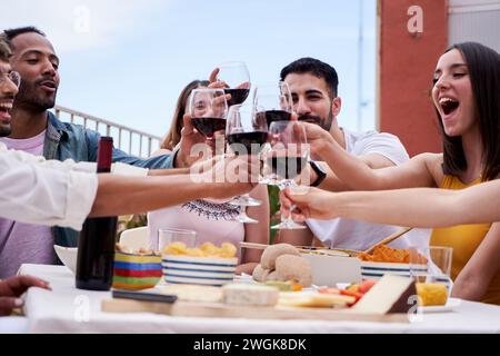 Gemischte Gruppe von Freunden, die gemeinsam auf einem Dach mit Rotwein feiern. Stockfoto