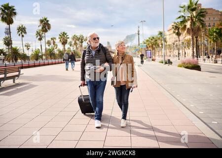 Lächelndes reifes kaukasisches Touristenpaar, das sich sorglos mit Gepäck an sonnigen Tagen im Freien umarmte. Stockfoto