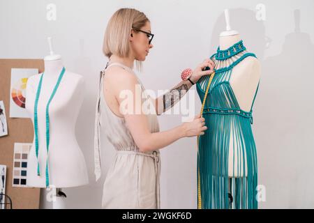 Schneiderin misst das Oberteil eines grünen Makramee-Kleides an der Schaufensterpuppe, die in einem Modeatelier arbeitet Stockfoto