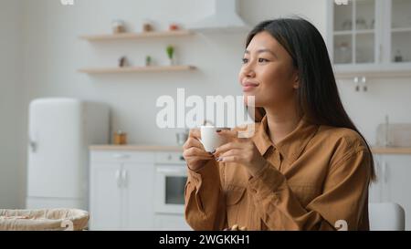 Entspanntes Mädchen asiatisch koreanische Frau in der Hausküche mit einer Tasse Kaffee trinken heißer Kräutertee multiethnische Dame nachdenklich ruhige Hausfrau Drink Stockfoto