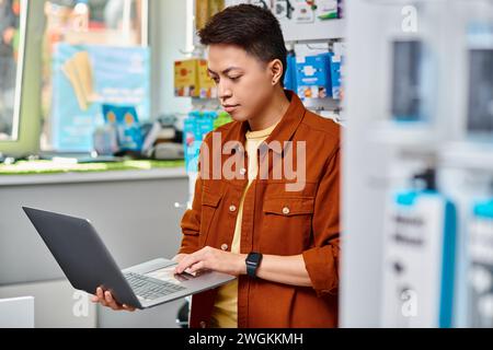 Konzentrierter asiatischer Mann, der Laptop benutzt, während er in einem privaten Elektronikgeschäft steht, einem kleinen Unternehmen Stockfoto