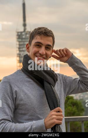 Junger braunhaariger Mann lächelt in die Kamera, während er sein Ohr während Sonnenuntergang berührt. Vertikal Stockfoto