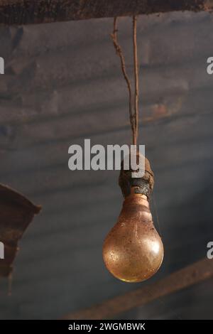 Nahaufnahme einer alten schmutzigen oder staubigen Glühlampe und hängend an einem Elektrokabel mit Metalldach und Rauch im Hintergrund, Dorfküchenlampe Stockfoto