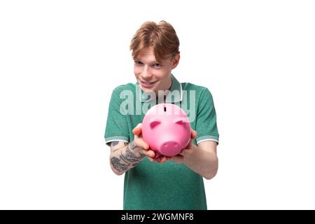 Ein junger Europäer mit roten Haaren hält eine rosa Sparkasse mit Geld und Münzen aus Stockfoto